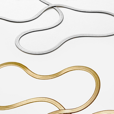 2 4mm herringbone necklaces of silver and 18k gold plated colors arranged in wave pattern lying on a white background showing the softness and flexible structure of the necklaces