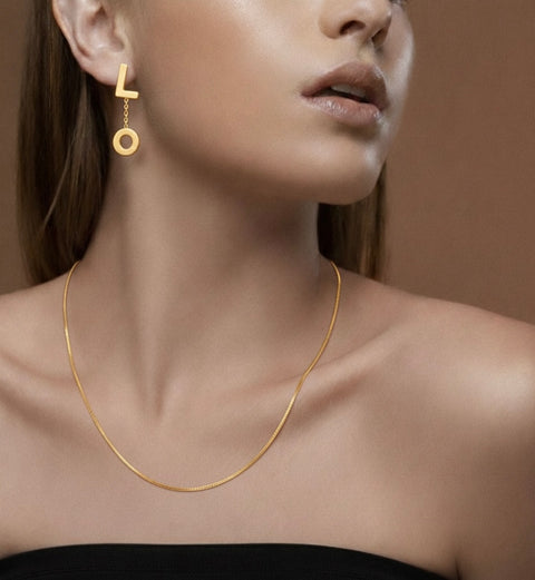 Close-up of a woman wearing B.Tiff ERloveG 18k gold plated drop earrings with linked letters "L" and "O" and a B.Tiff C002G 18K gold plated necklace against a brown background