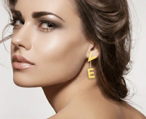 Close-up of a woman wearing B.Tiff ERloveG gold plated drop earrings with linked letters 'V' and 'E' on a neutral background