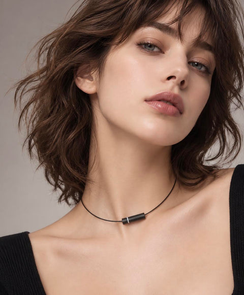 A close-up of a woman with short brown hair wearing a B.TIff PT080B minimalist black cord necklace with a dark metallic cylindrical pendant, set against a neutral gray background.