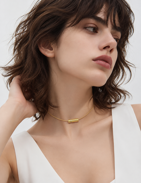 A close-up of a woman wearing a delicate B.Tiff PT080G gold chain necklace with a small horizontal bar pendant, styled with a white sleeveless top against a clean white background.