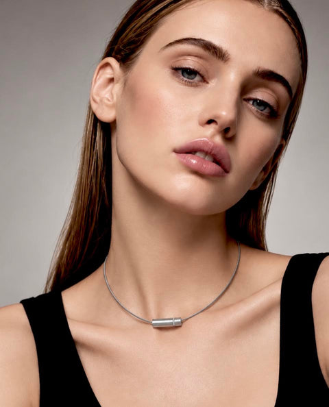 A close-up of a woman wearing a B.TIff PT080W minimalist silver cylindrical pendant necklace on a thin chain, paired with a black top against a neutral gray background.