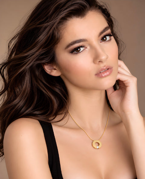 A woman with long wavy brown hair models a delicate B.Tiff PT100G gold chain necklace featuring a small gold ring pendant with a clear central gemstone, set against a solid warm beige background.