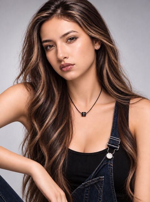 A woman with long, wavy brown hair wearing a black tank top and blue denim overalls, featuring a B.Tiff PT118B black cord necklace with a small black barrel pendant paved with diamonds. She is posed against a solid gray studio background.