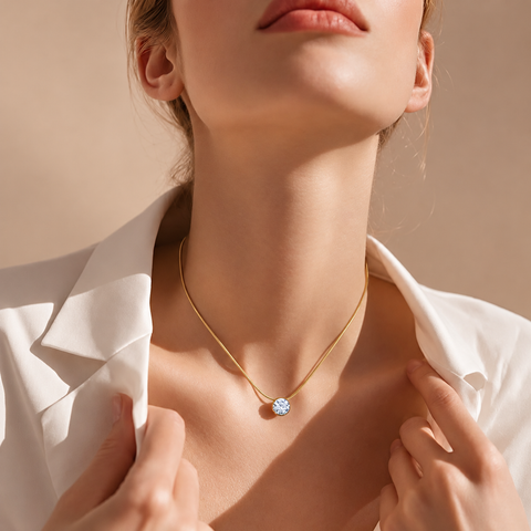 A close-up lifestyle shot of a model wearing a delicate gold chain necklace with a B.Tiff PT201G 2 carat, clear, round-cut gemstone pendant. The model is wearing a white collared shirt against a warm beige background.