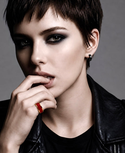 A close-up portrait of a model with a short dark haircut, heavy black eye makeup, and a black leather jacket, wearing a B.Tiff RG800RGR bold red ring on one finger against a neutral gray background.