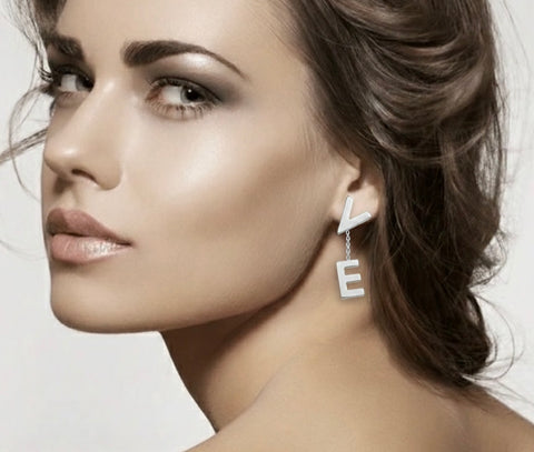 Close-up of a woman wearing B.Tiff ERloveW silver 'LOVE' earrings with linked letters 'V' and 'E' charms on a neutral background