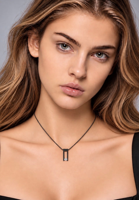 A close-up portrait of a woman wearing a delicate B.Tiff PT089B black cord necklace with a small black rectangular pendant, set against a neutral gray background.