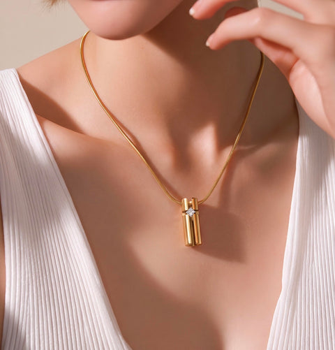 A close-up lifestyle shot of a model wearing a B.Tiff PT099G gold pendant necklace. The necklace features a polished gold cylinder pendant with a small clear gemstone accent, suspended from a delicate gold chain.