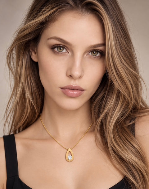 A close-up portrait of a woman with long highlighted brown hair wearing a delicate B.Tiff PT106G gold pendant necklace with a teardrop-shaped clear gemstone. She is wearing a black top against a soft, neutral beige background.