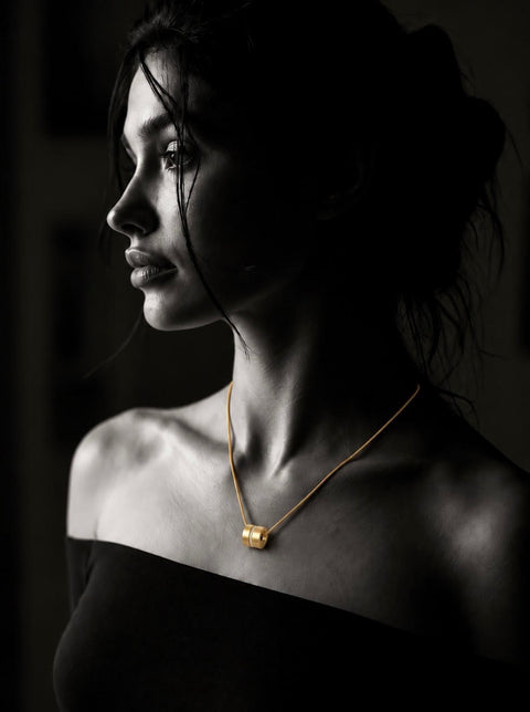 A woman in profile wearing a delicate B.Tiff PT118G gold chain necklace with a small geometric barrel pendant paved with diamonds, shown in dramatic high-contrast black and white lighting.