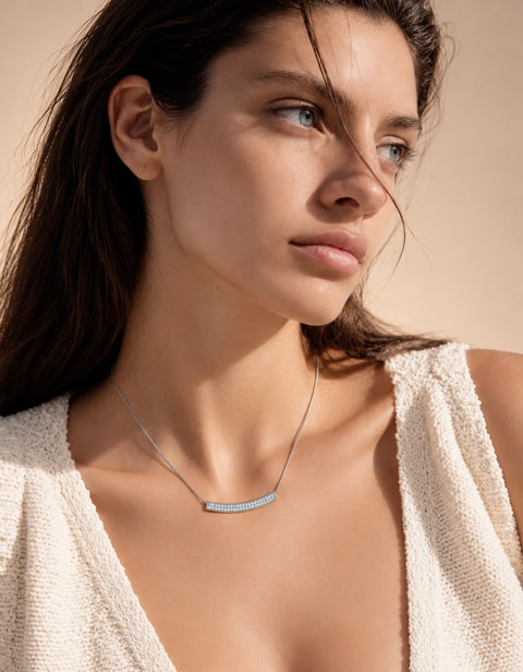A close-up of a woman wearing a delicate B.Tiff PT222W silver necklace with a curved bar set with small clear stones, paired with a textured cream knit top against a warm beige background.